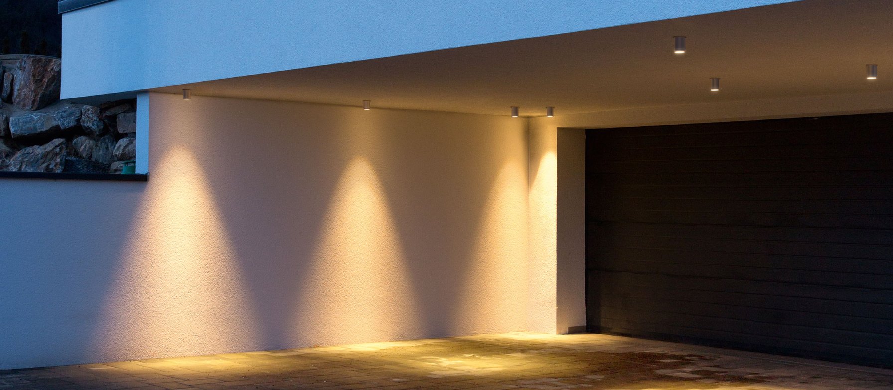 Carport eines modernen Wohnhauses zur blauen Stunde, Deckenspots werfen warme Lichtkegel auf die Wand