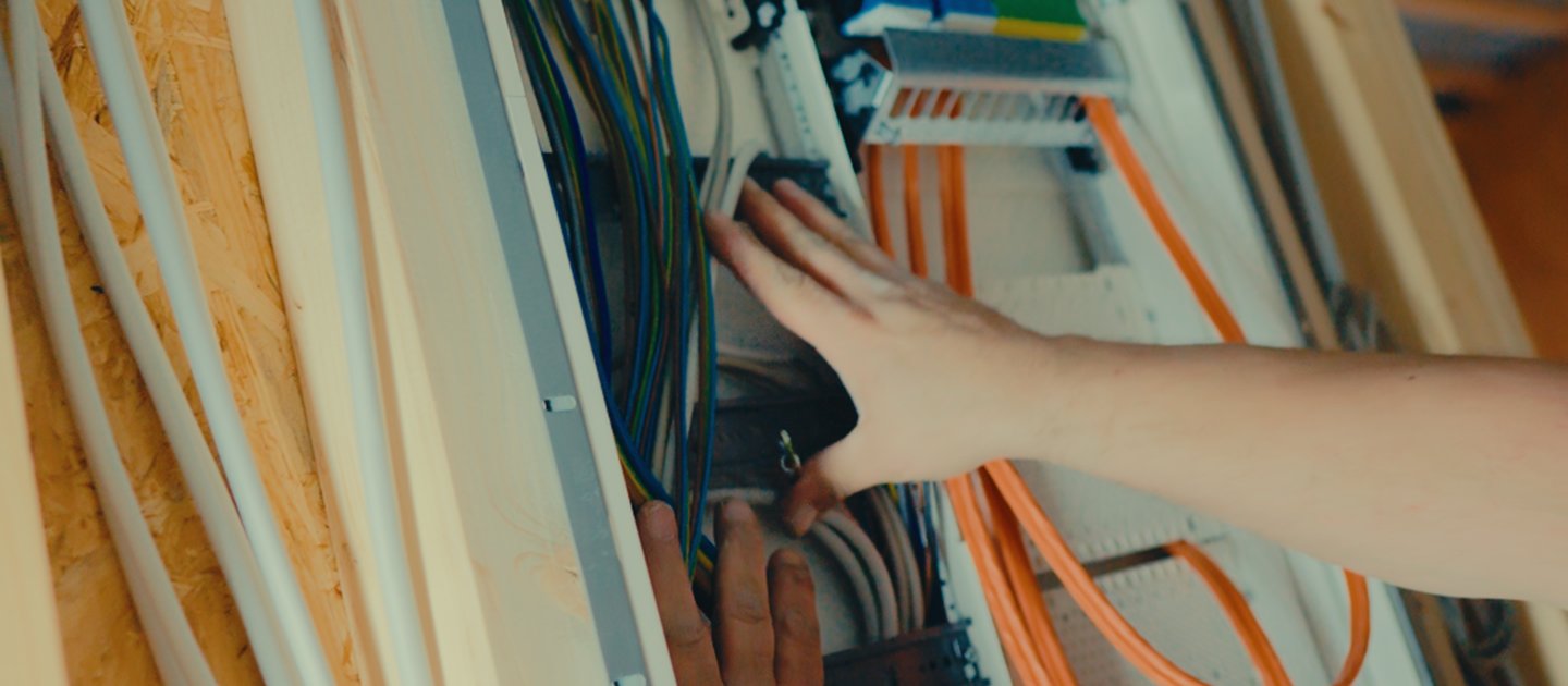 Zwei Hände verdrahten eine offene Unterverteilung in einer Holzständerwand, daneben oranges Datenkabel