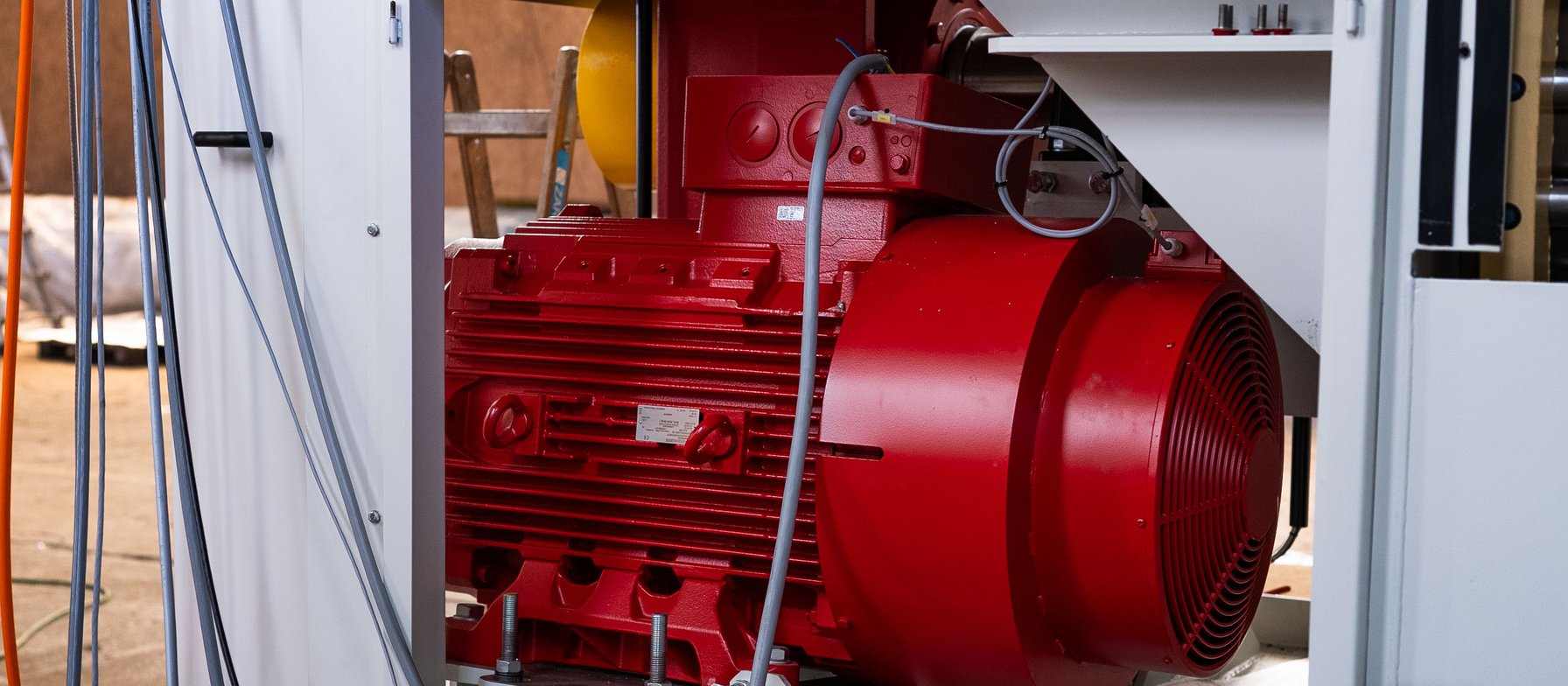 Großer roter Elektromotor in einer Maschinenverkleidung mit Lüftungsgitter und Kabelbündeln